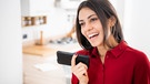 Eine junge, gut gelaunte Frau mit roter Bluse spricht eine Sprachnachricht in ihr Smartphone ein | Bild: mauritius-imeages